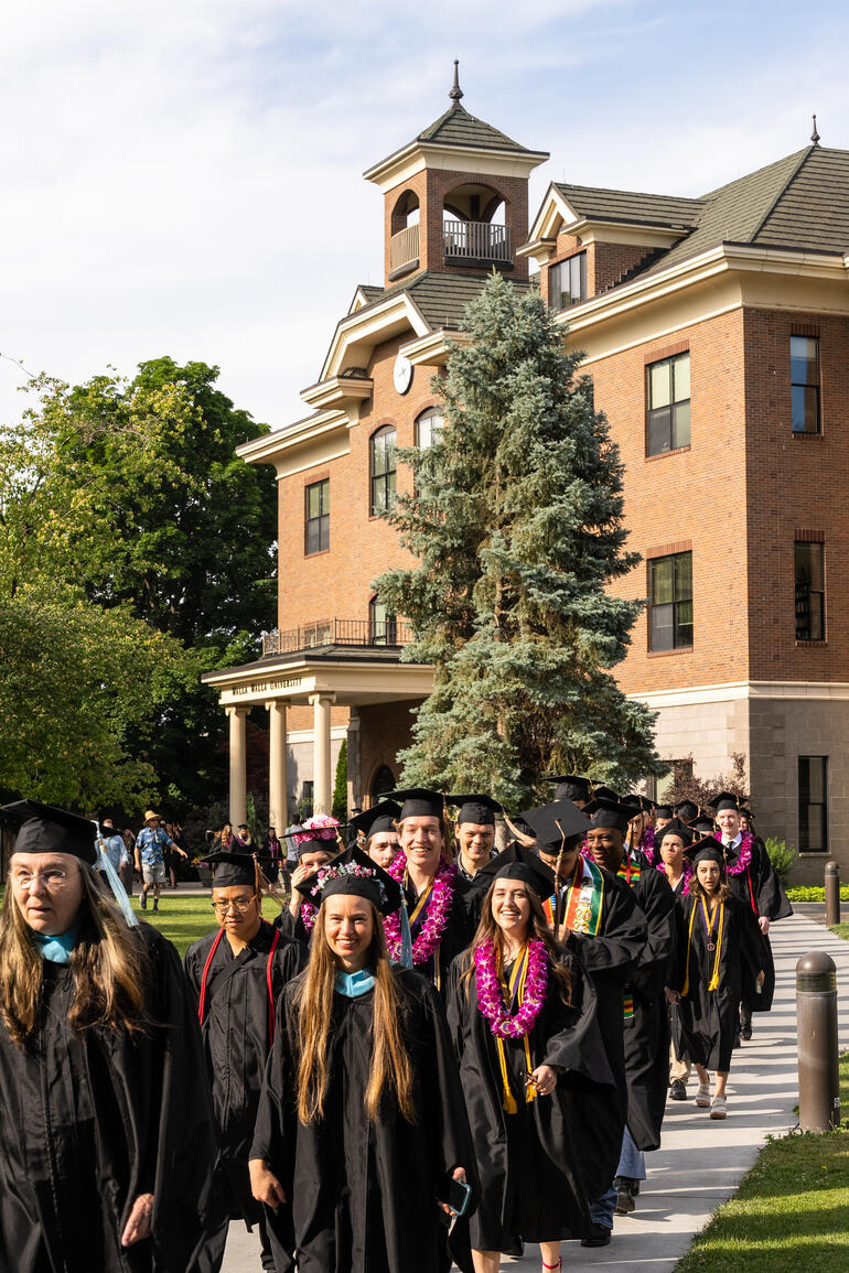 Many Walla Walla University graduates in regalia march along sidewalk in front of administration building.