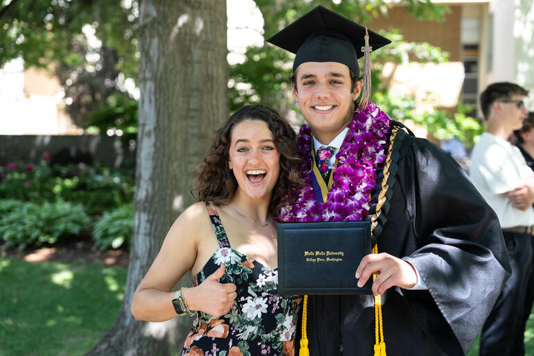 Woman stands next to male graduate holding diploma and smiling.