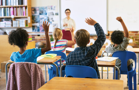 Classroom of children with hands raised.