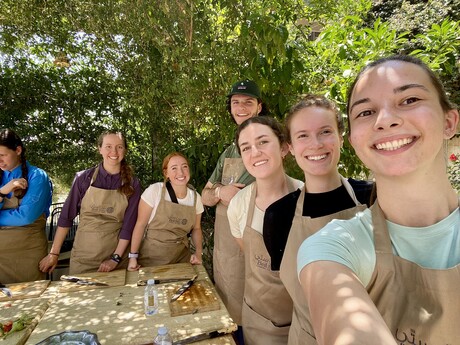 Students wearing aprons gather around outdoor food prep station.