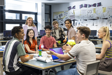 Wide shot of a group of students and their teacher gathered around a table in a technology classroom.