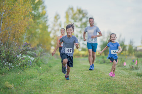 A small young mixed race family is seen participating in a race together as they run outdoors on a warm fall day.  They are each dressed comfortably in shorts and t-shirts and have race bibs on.