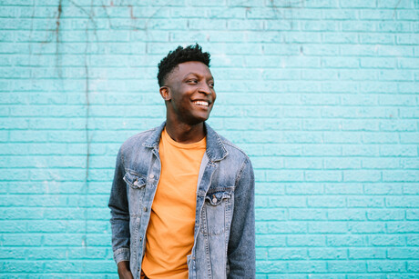 A smiling African American man stands in front of a blue wall wearing nice casual clothing. 