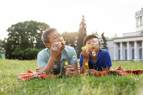 asian old father together with son lie on the grass in the park and blow soap bubbles and rejoice in summer, korean boy rest and play with dad outdoors, concept of parental love and care