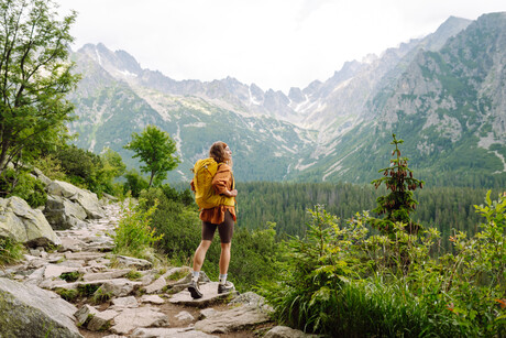 Happy woman with a yellow hiking backpack enjoying the mountain landscape. A young traveler travels along mountain paths. Adventure, travel concept.