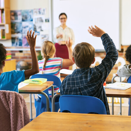 Classroom of children with hands raised.