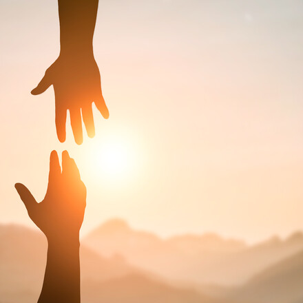 Silhouette of two people hands reaching to each other for help in sunset sky and orange sun. Friendship, teamwork, help, faith and hope concept.