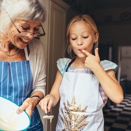Senior woman in apron making batter for cake. Little girl tasting cake batter standing in kitchen with grandmother.