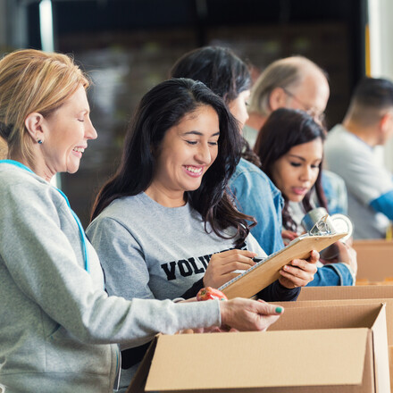 Mature adult Caucasian woman is standing with mid adult Hispanic woman as they pack cardboard boxees full of donated food in charity food bank. Other volunteers are lined up behind them, also sorting donated groceries into boxes. Hispanic woman is writing on checklist on clipboard.