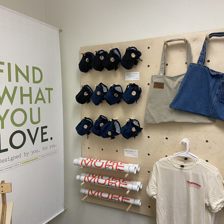 Tshirts, tote bags, posters, and chalk bags hang on wooden display next to Well-Designed Goods store sign.