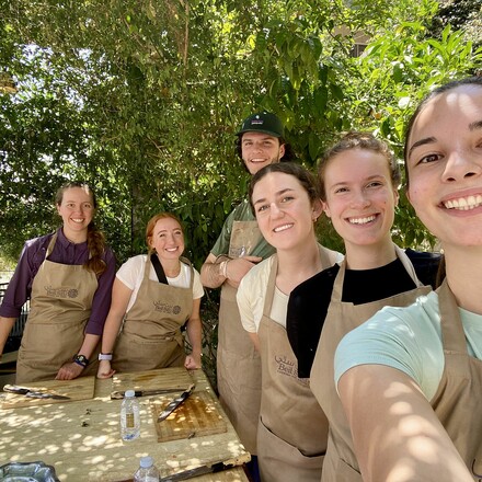 Students wearing aprons gather around outdoor food prep station.