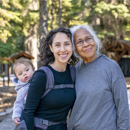 A young mixed race woman carries her one year old daughter on her back in an ergonomic baby carrier while out on a hike with her senior mother. The multi-generation family is staying active and healthy outdoors in Oregon.