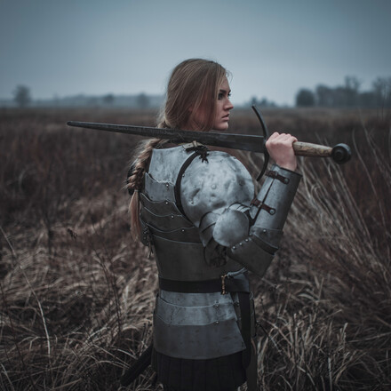 Girl in image of Jeanne d'Arc in armor and with sword in her hands stands on meadow in middle of dry grass. Back view.