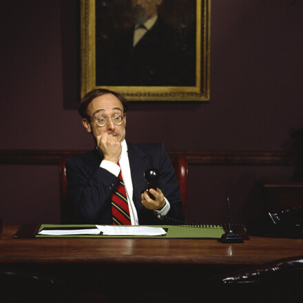 Businessman sits at a desk while holding a phone in shock.
