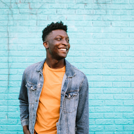 A smiling African American man stands in front of a blue wall wearing nice casual clothing.