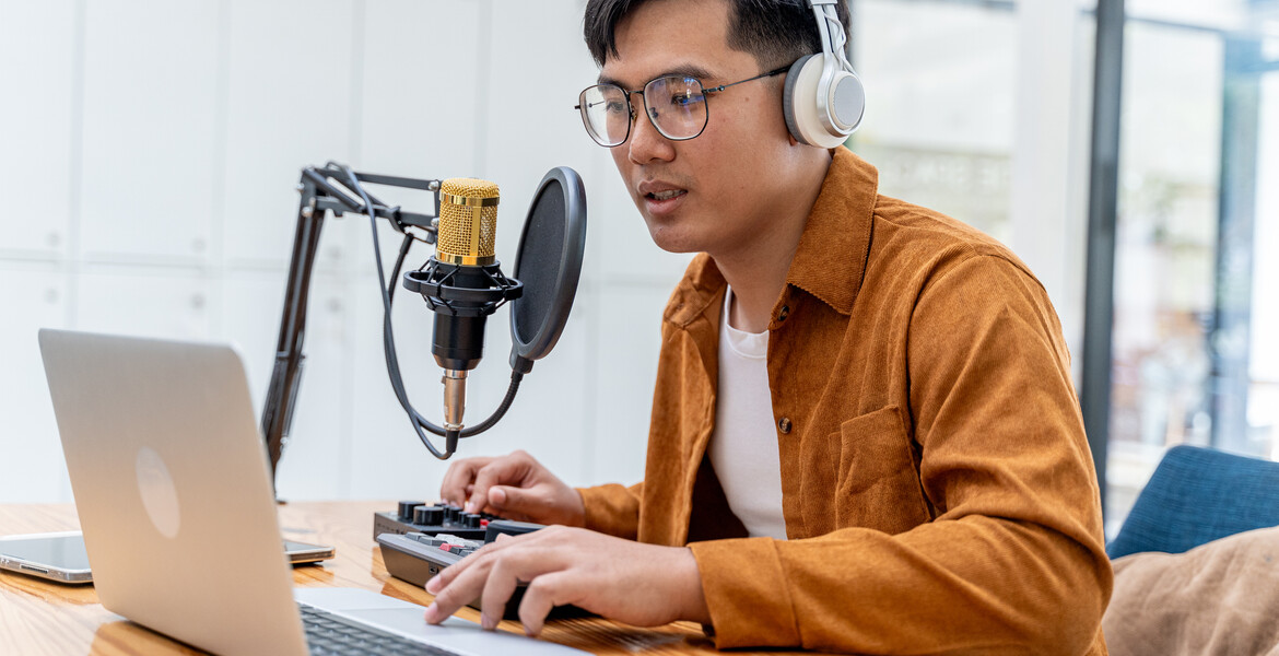 Young Asian man speaking to microphone during his live podcast while checking the feedback from his online audiences from his laptop computer