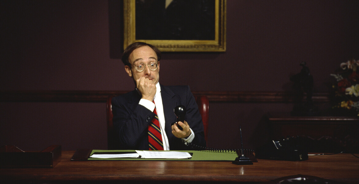 Businessman sits at a desk while holding a phone in shock.