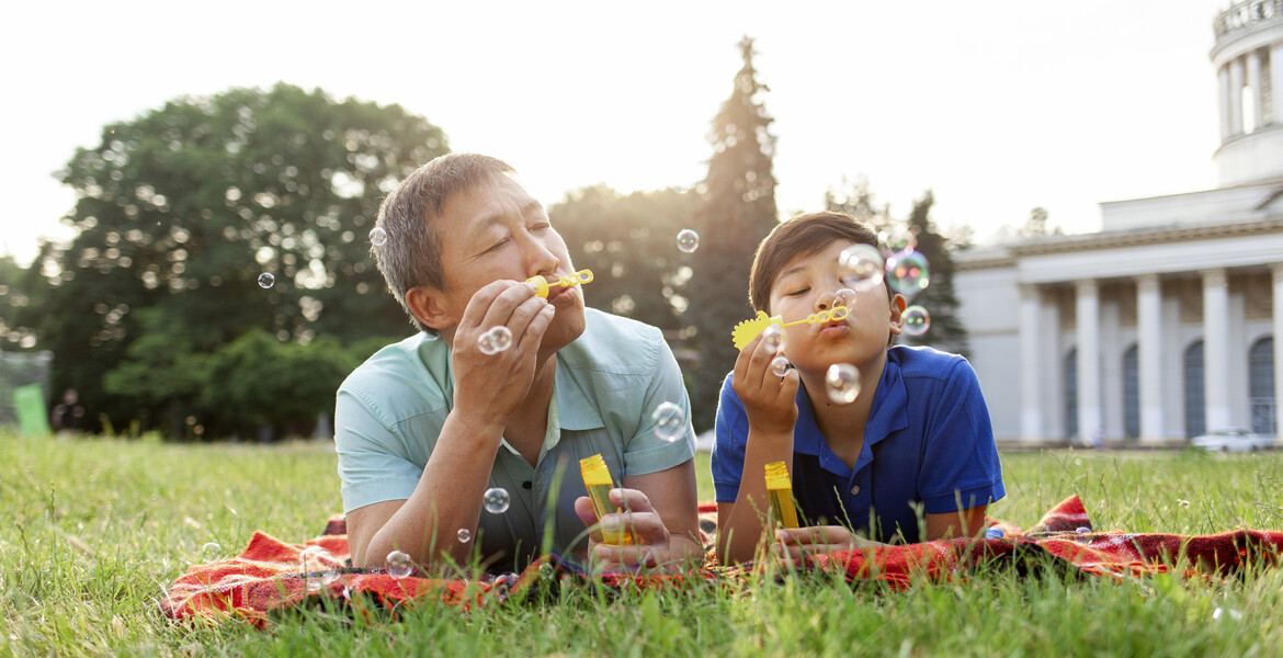 asian old father together with son lie on the grass in the park and blow soap bubbles and rejoice in summer, korean boy rest and play with dad outdoors, concept of parental love and care